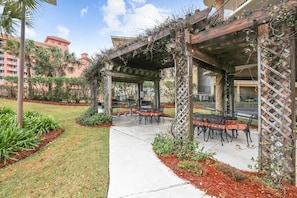An outdoor patio with a pergola, tables, and chairs, surrounded by landscaped gardens and a building in the background.