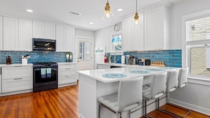 A modern kitchen with white cabinets, a central island with bar stools, a built-in oven, and a blue-tiled backsplash.