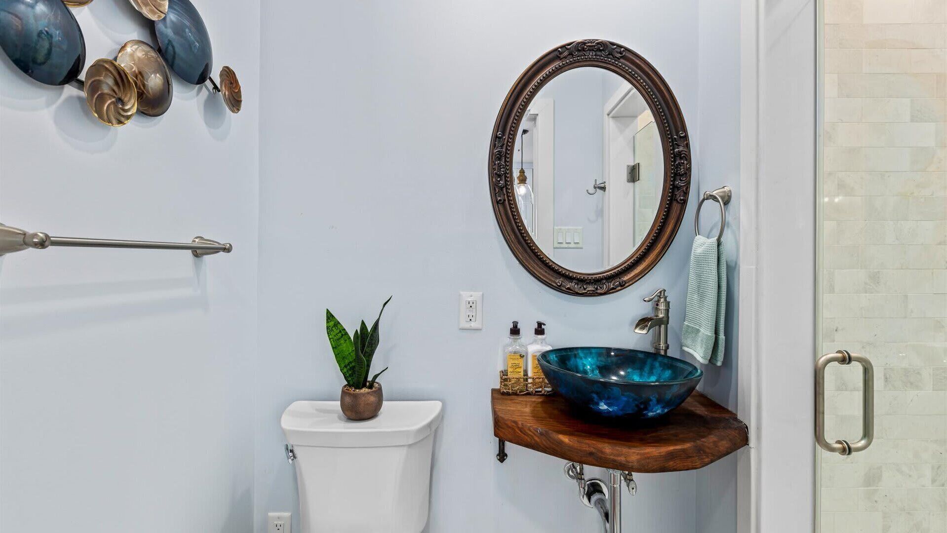 Ein Badezimmer mit einer weißen Toilette, einem hölzernen Waschtisch mit einer blauen Glas-Schüssel-Spüle, einem Spiegel und einer Pflanze auf der Toilette.