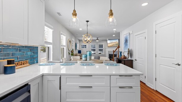 A modern kitchen with white cabinetry, a central island, pendant lighting, and a view to the living area.