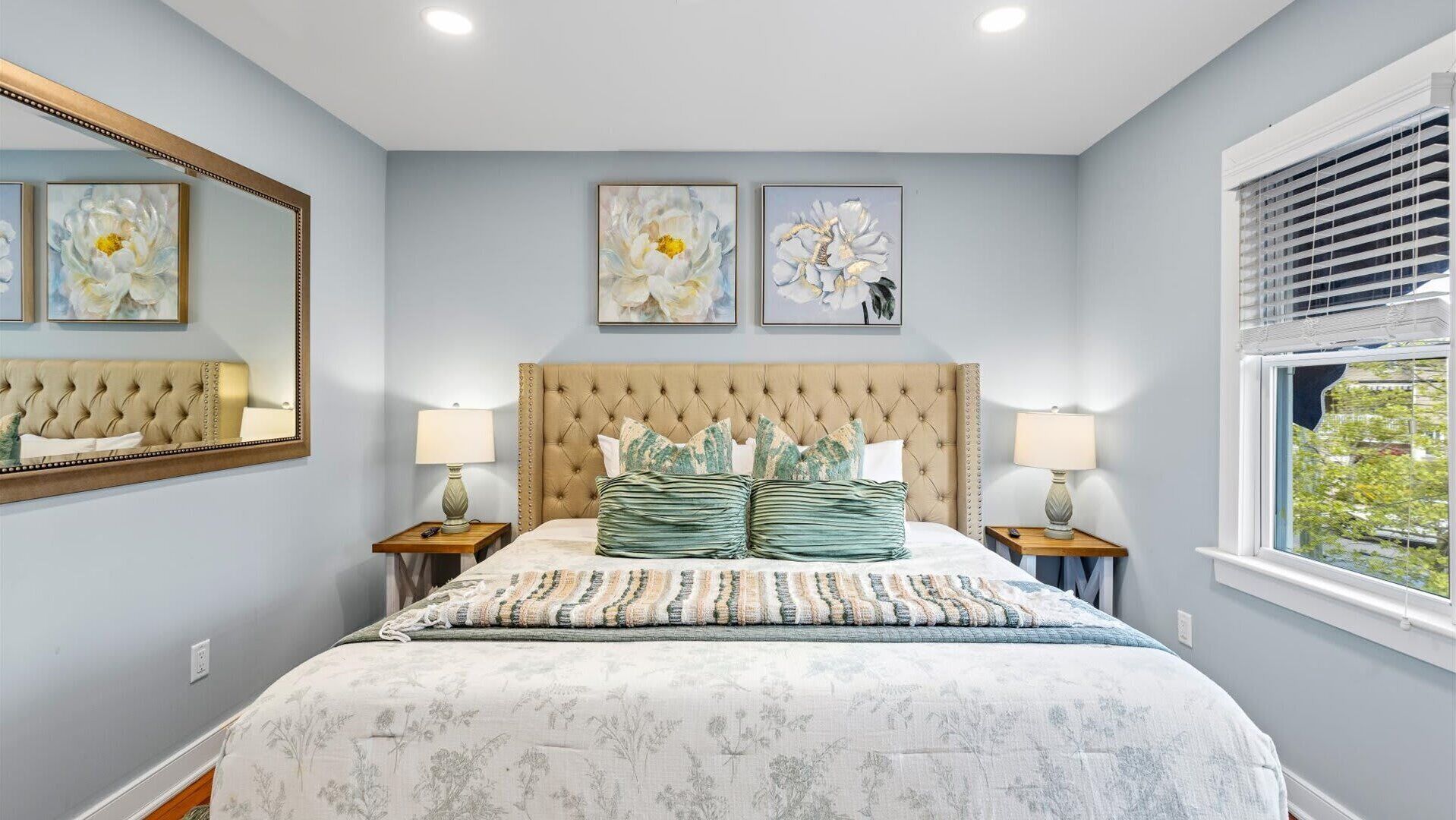 Un dormitorio con cabecera capitonada, ropa de cama estampada con motivos florales, dos lámparas de noche y un ventanal con persianas.