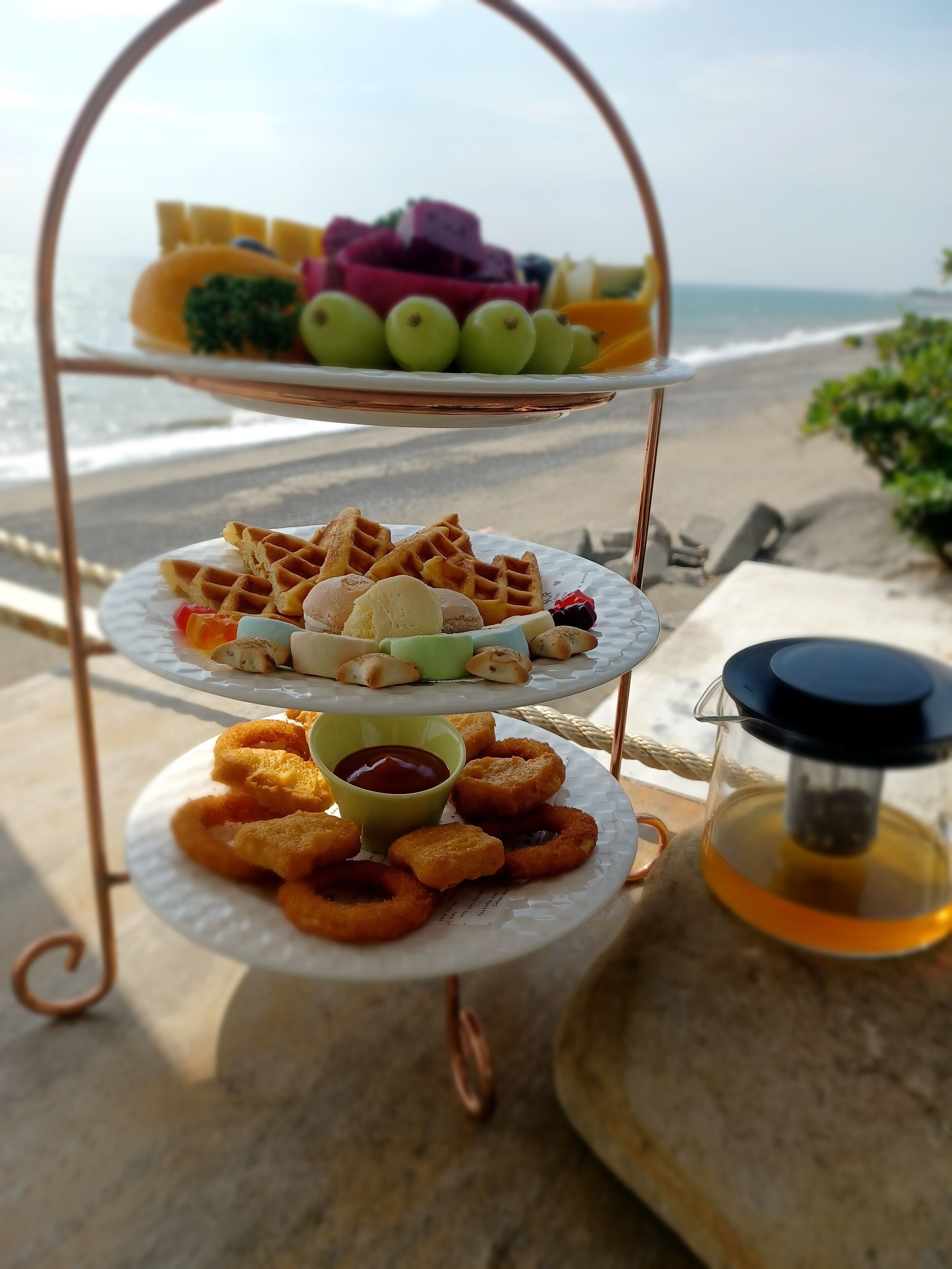Una bandeja de tres niveles con diversos bocadillos y una bebida sobre una mesa cerca de la playa.