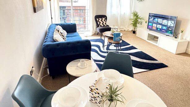 Ein modernes Wohnzimmer mit einem blauen Sofa, einem Esstisch mit weißen Tellern, einem Fernseher und einem großen Fenster mit Vorhängen.