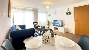 Ein Wohnzimmer mit einem blauen Sofa, einem Esstisch mit weißen Tellern und Schüsseln, einem Fernseher auf einem Schrank und einer Tür.