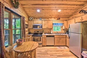 A wooden cabin kitchen with a wooden table, a stove, a microwave, a refrigerator, and a window with curtains. - Hot Tub, Firepit, Minutes to Helen (Clarkesville)