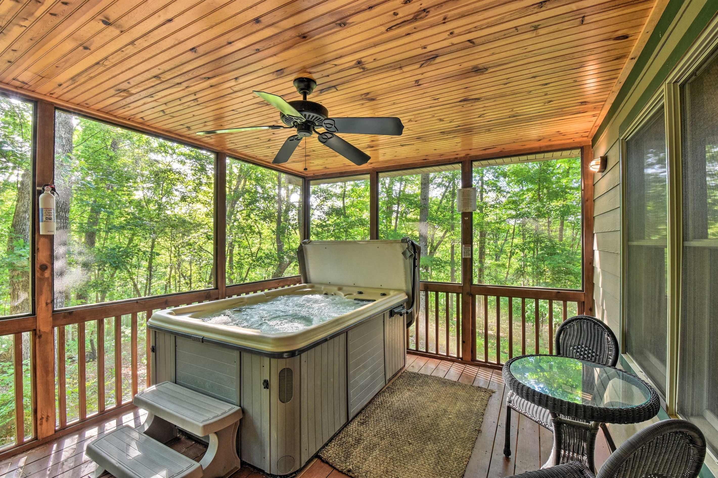 A hot tub on a wooden deck with a ceiling fan, surrounded by glass windows offering a forest view.