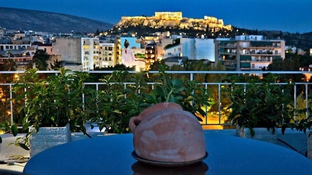 Una mesa con una jarra de cerámica y una carta de juego en un balcón con vistas a un paisaje urbano que incluye una colina destacada y un edificio iluminado.