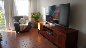 A living room with a sofa, a TV on a wooden stand, a potted plant, and a sliding glass door. - Bongaree Getaway, Clean Comfy, and Homely. (Bongaree)
