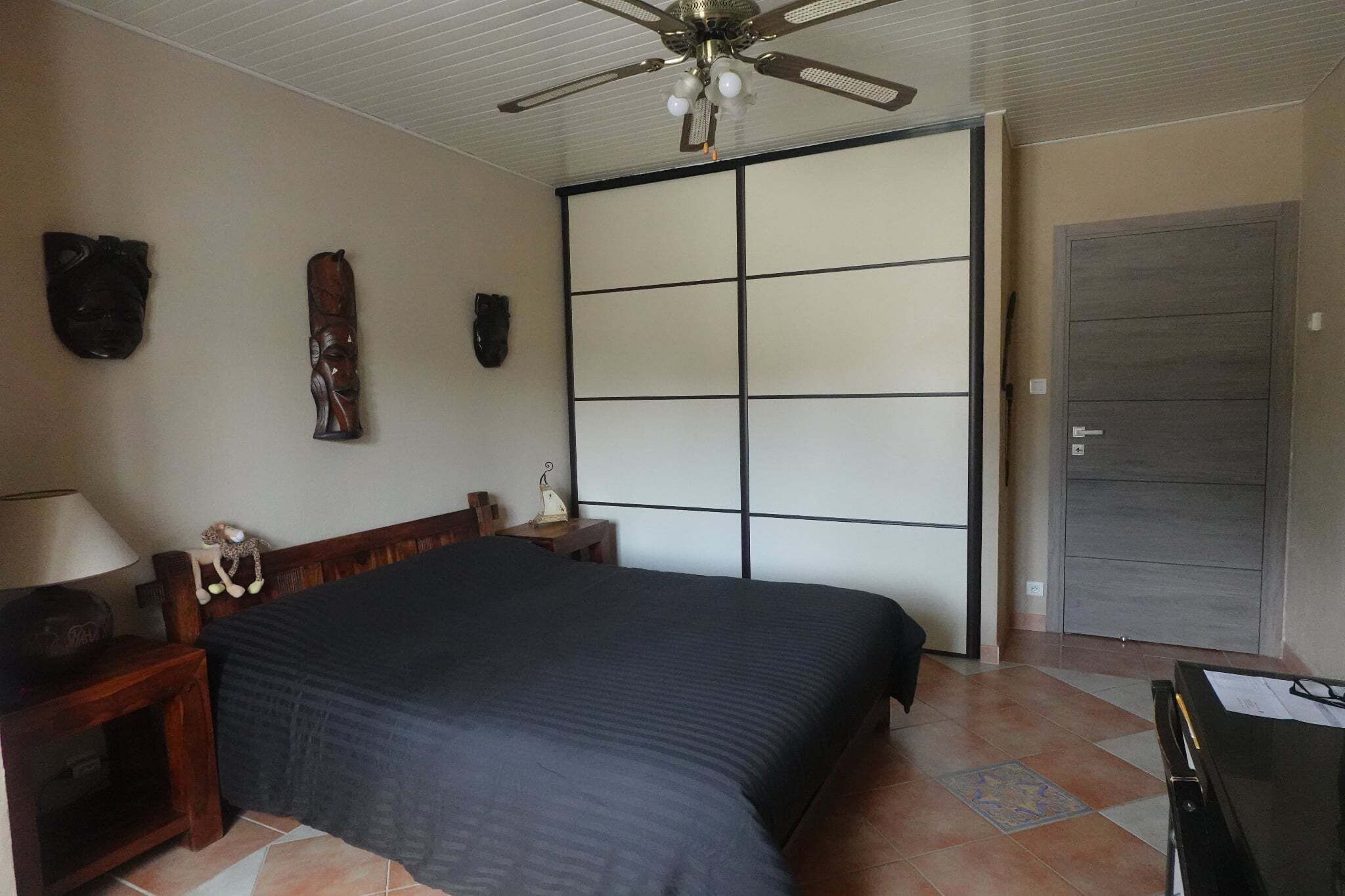 Une chambre à coucher comprenant un lit, un ventilateur de plafond, une armoire et un bureau avec une lampe.