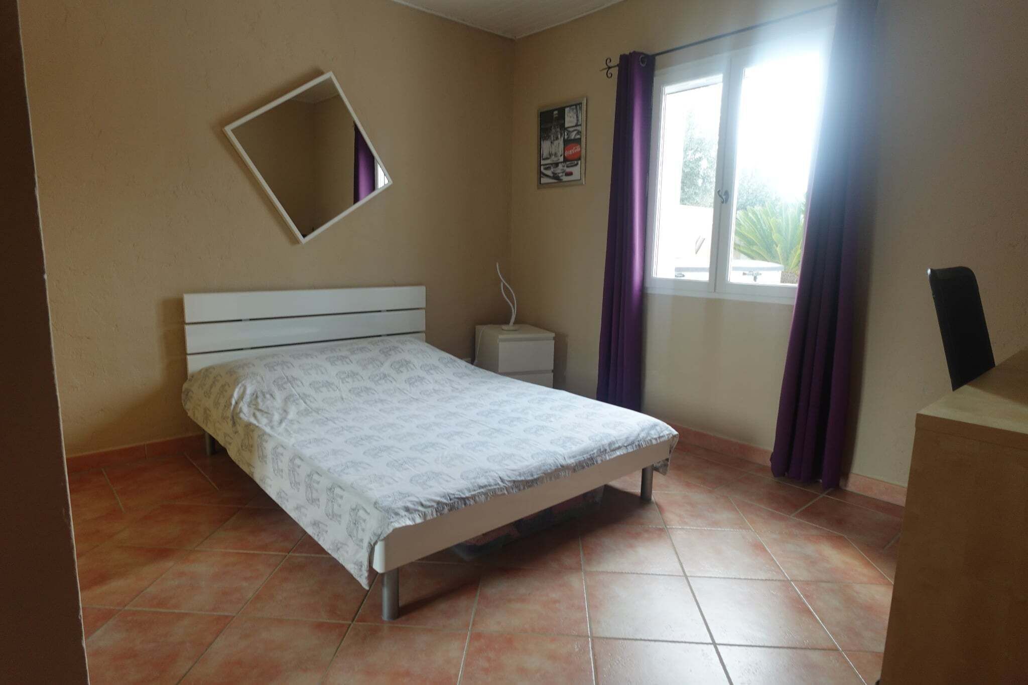 Ein Schlafzimmer mit Bett, Nachttisch, Spiegel und einem Fenster mit Vorhängen.