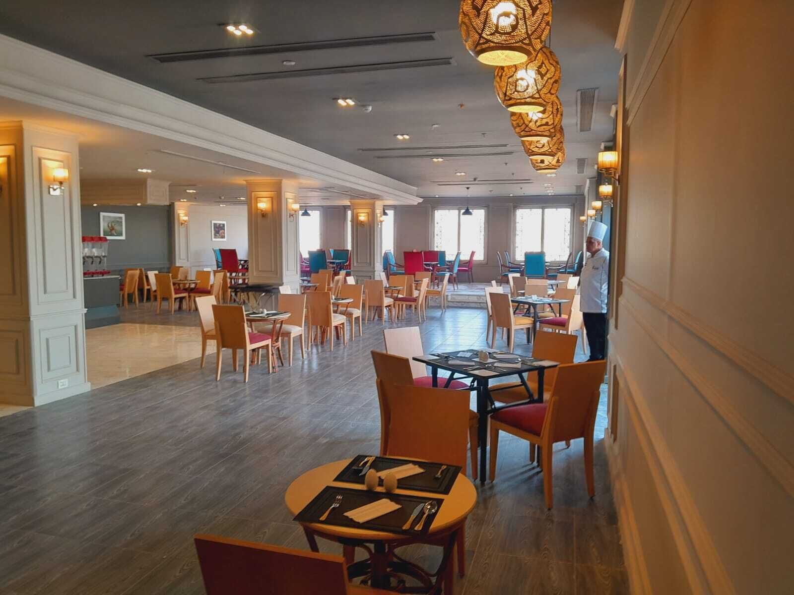 Ruime hotel eetzaal met meerdere tafels en stoelen, een keuken op de achtergrond en een kok in koksoutfit.
