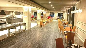 A hotel dining area with multiple tables and chairs, a buffet setup on the left, and large windows along the back wall.