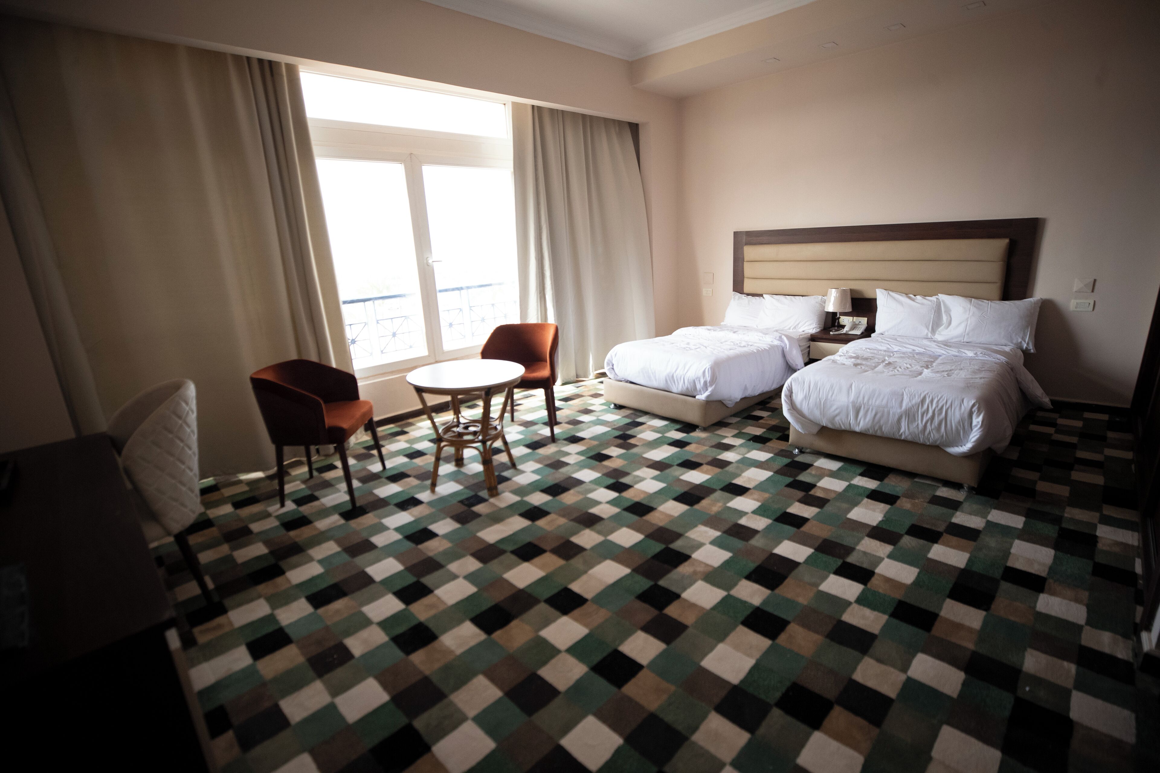 A hotel room with a checkered floor, two beds, a round table, and a chair.