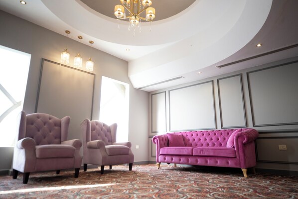 Una habitación con dos sillones, un sofá rosa, alfombra estampada, un candelabro y ventanas amplias.