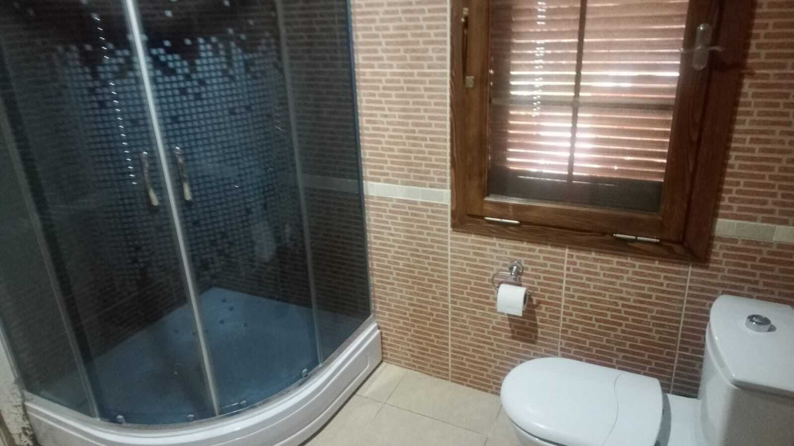 A bathroom with a glass shower enclosure, a toilet, and a window with blinds.