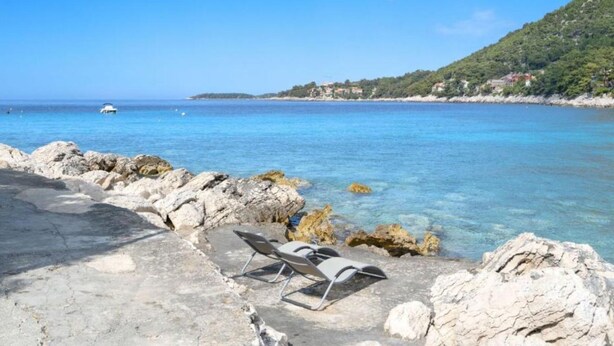 A rocky beach with clear blue water, a few lounge chairs, and a distant view of a hilly coastline with buildings. - Holiday Home Seafront Oleandar (Blato)