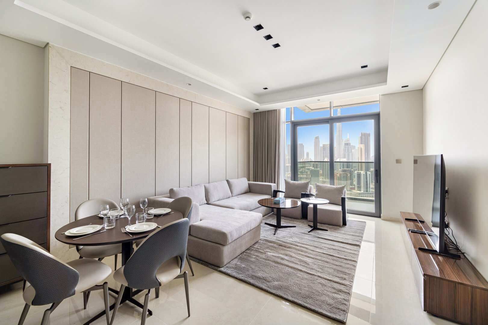 Une chambre d’hôtel moderne dotée d’un coin repas, d’un canapé, de deux petites tables, d’une télévision et offrant une vue sur la ville.