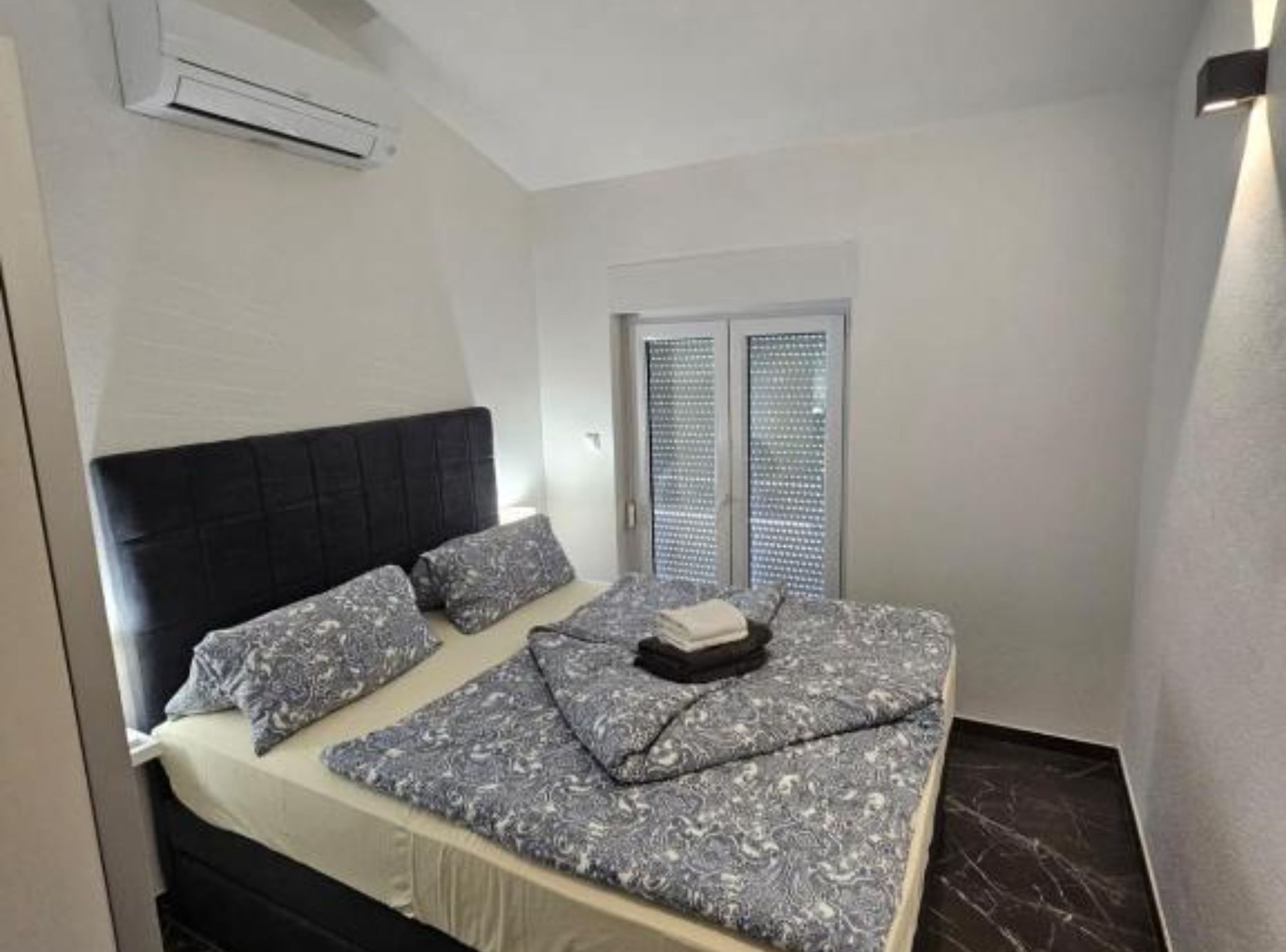 A small, modern bedroom with a bed, a window with blinds, and an air conditioning unit.