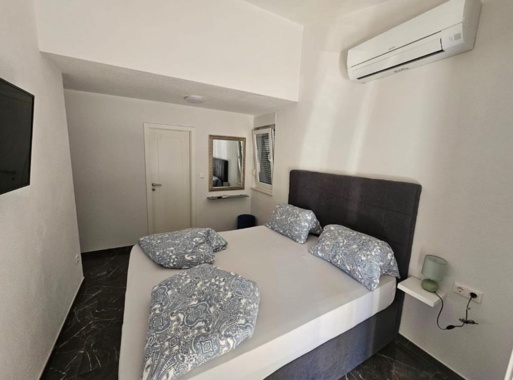 A bedroom with a bed, two pillows, a bedside table, a mirror, an air conditioning unit, and a door.