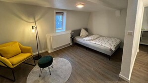 A small, well-lit room with a single bed, a yellow chair, a round table, and a window. - Apartment in Vallendar Near Rhine Promenade (Vallendar)