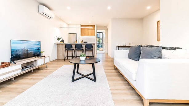 Un salon moderne avec un téléviseur à écran plat, un canapé blanc, un coin repas avec des chaises et une cuisine en arrière-plan.