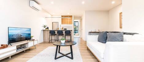 Een moderne woonkamer met een flatscreen televisie, een witte bank, een eethoek met stoelen en een keuken op de achtergrond.