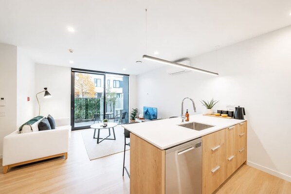 A modern kitchen with a large island, wooden cabinets, and a dishwasher. There is a dining area with a table and chairs, and a sofa.