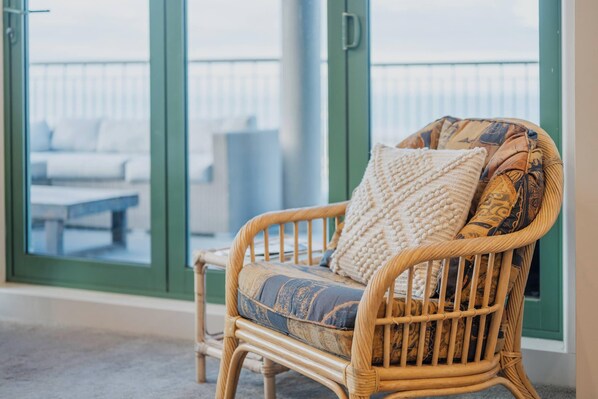 Una silla de mimbre con cojines estampados en una habitación con ventanas amplias y vistas a muebles de exterior.