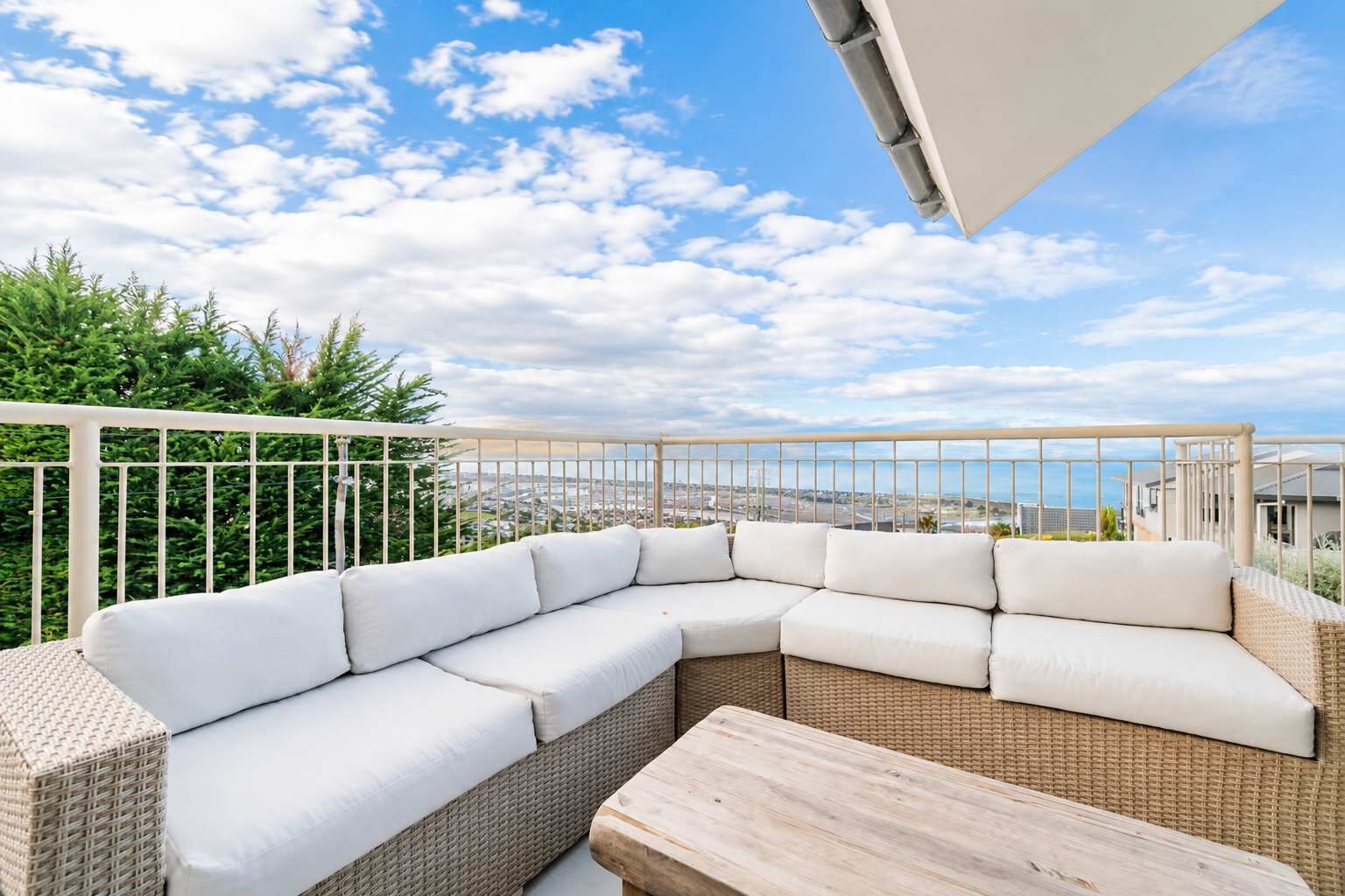 A balcony with a wicker sofa and a wooden table, overlooking a cityscape.