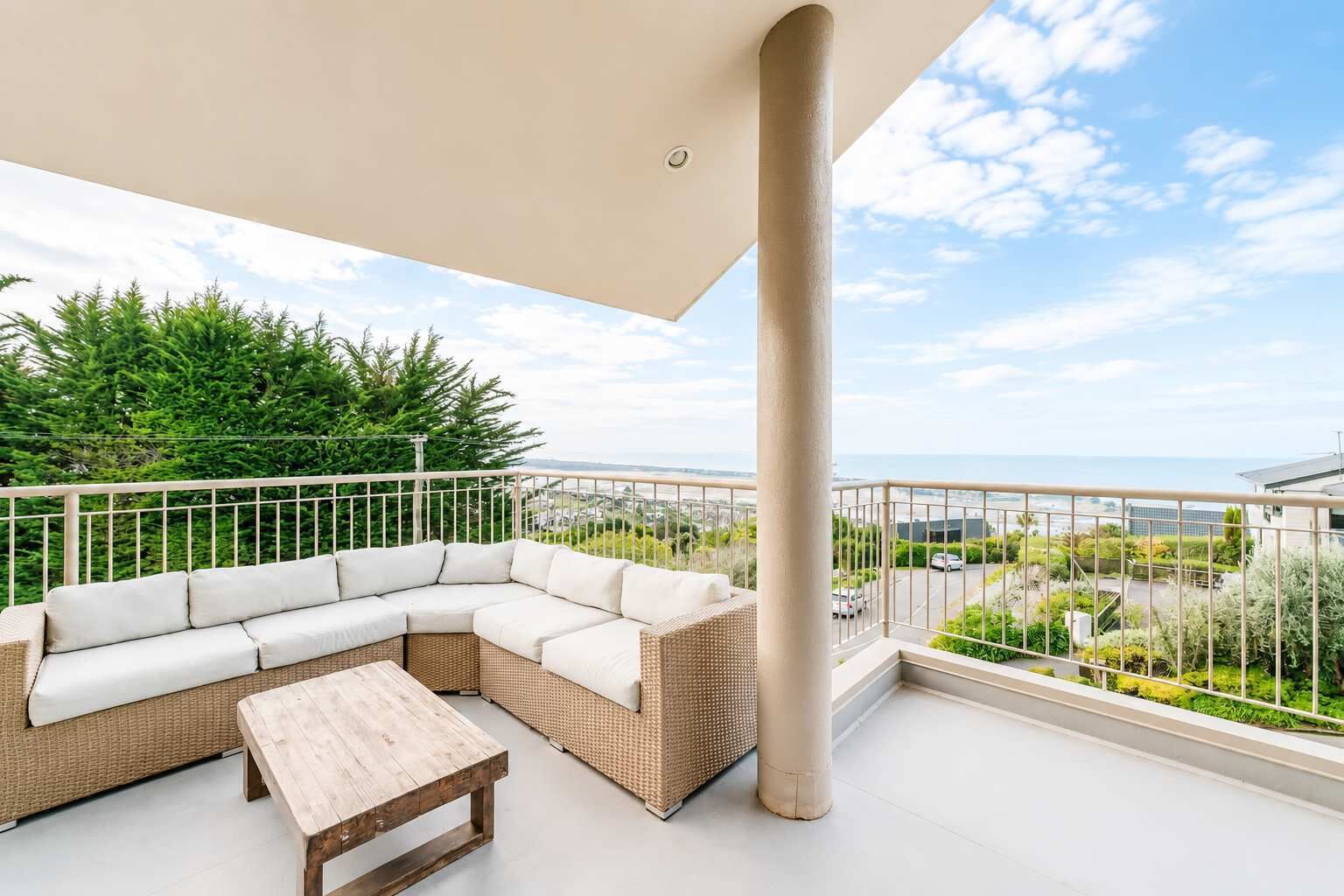 A balcony with a white sectional sofa and a wooden table, overlooking a cityscape and greenery.