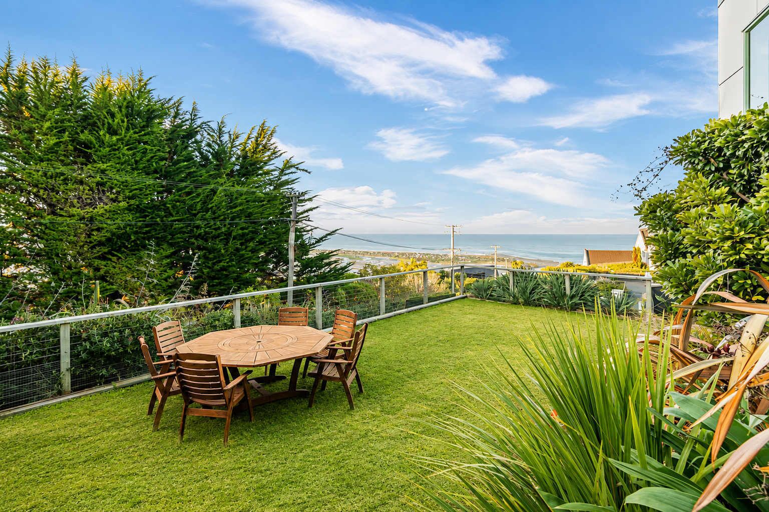 A fenced outdoor area with a round wooden table and chairs, surrounded by green grass and various plants.