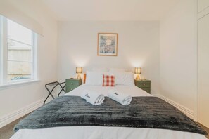 Ein ordentlich bezogenes Bett mit weißen Laken und einer schwarzen Decke, zwei Nachttische mit Lampen, ein kleiner, grüner Beistelltisch und ein Fenster mit Blick auf einen Zaun.