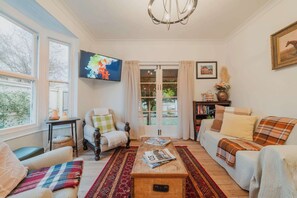 Ein gemütliches Wohnzimmer mit Sofa, Sessel und Couchtisch. Ein Fernseher ist an der Wand befestigt, und es gibt ein Bücherregal mit Büchern und Dekoartikeln.