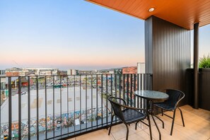 A balcony with a view of a parking lot and buildings, two chairs, and a small table.