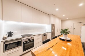 A modern kitchen with white cabinetry, a stainless steel refrigerator, a built-in oven, and a wooden dining table with a potted plant.