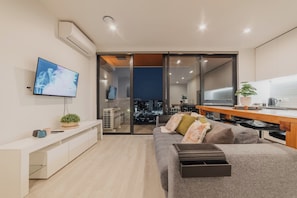 Un salon moderne avec un canapé gris, un téléviseur à  écran plat, une table à manger en bois et une porte-fenêtre coulissante donnant sur un balcon avec vue sur la ville.
