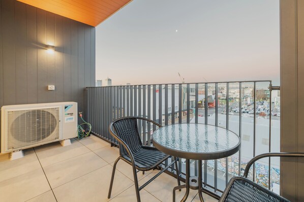 A balcony with a round glass table and two wicker chairs, overlooking a cityscape with buildings and a busy street.