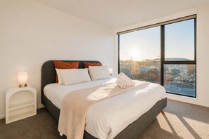 A bedroom with a large window, a bed with pillows, a bedside table with a lamp, and a view of a cityscape.