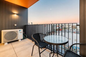 Een balkon met een ronde glazen tafel en twee stoelen, een airconditioning en uitzicht op een stadsgezicht.