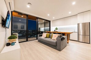 A modern living room with a grey sofa, a wooden dining table, and a kitchen area with white cabinets and a stainless steel refrigerator.