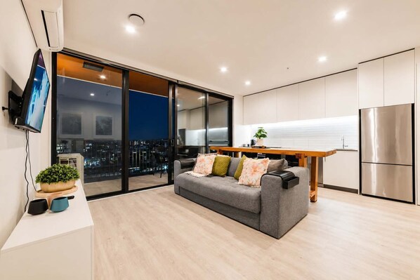 A modern living room with a grey sofa, a wooden dining table, and a kitchen area with white cabinets and a stainless steel refrigerator.