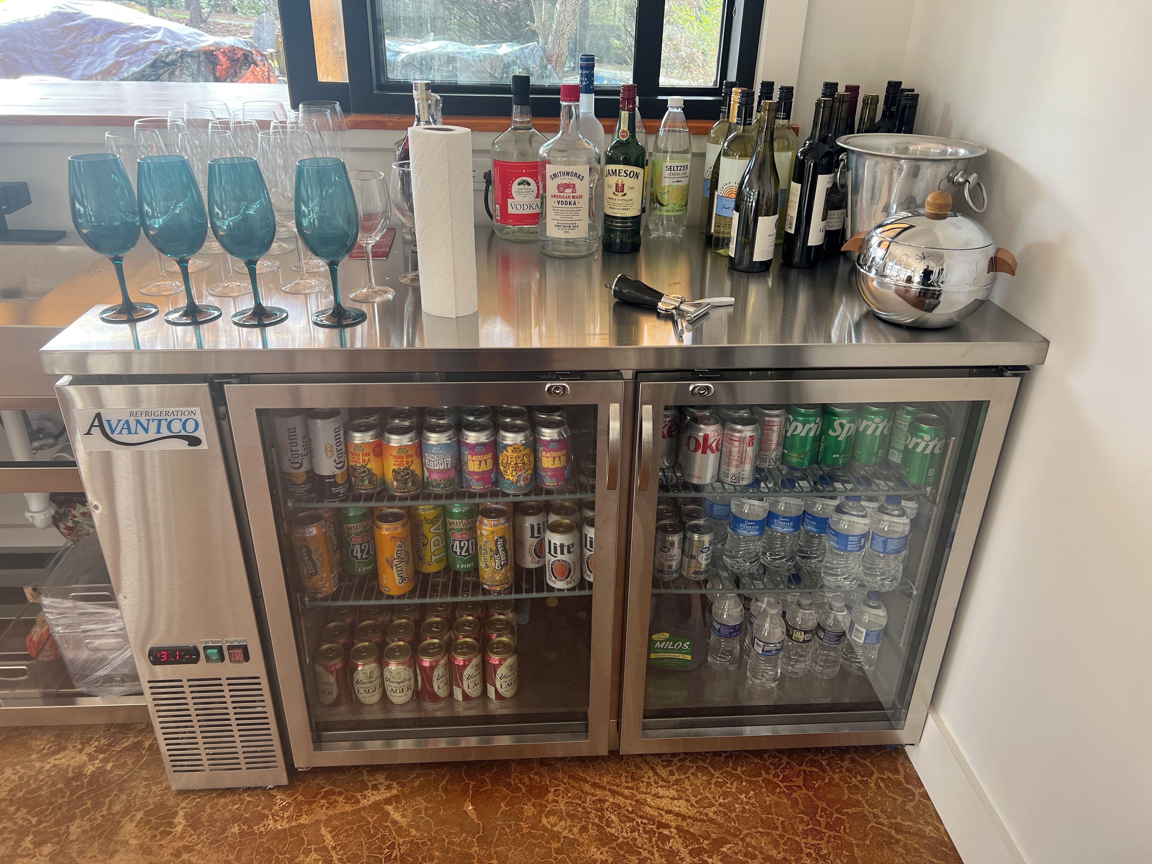 A bar area with a refrigerator stocked with beverages, a selection of bottles, and glasses on the counter.