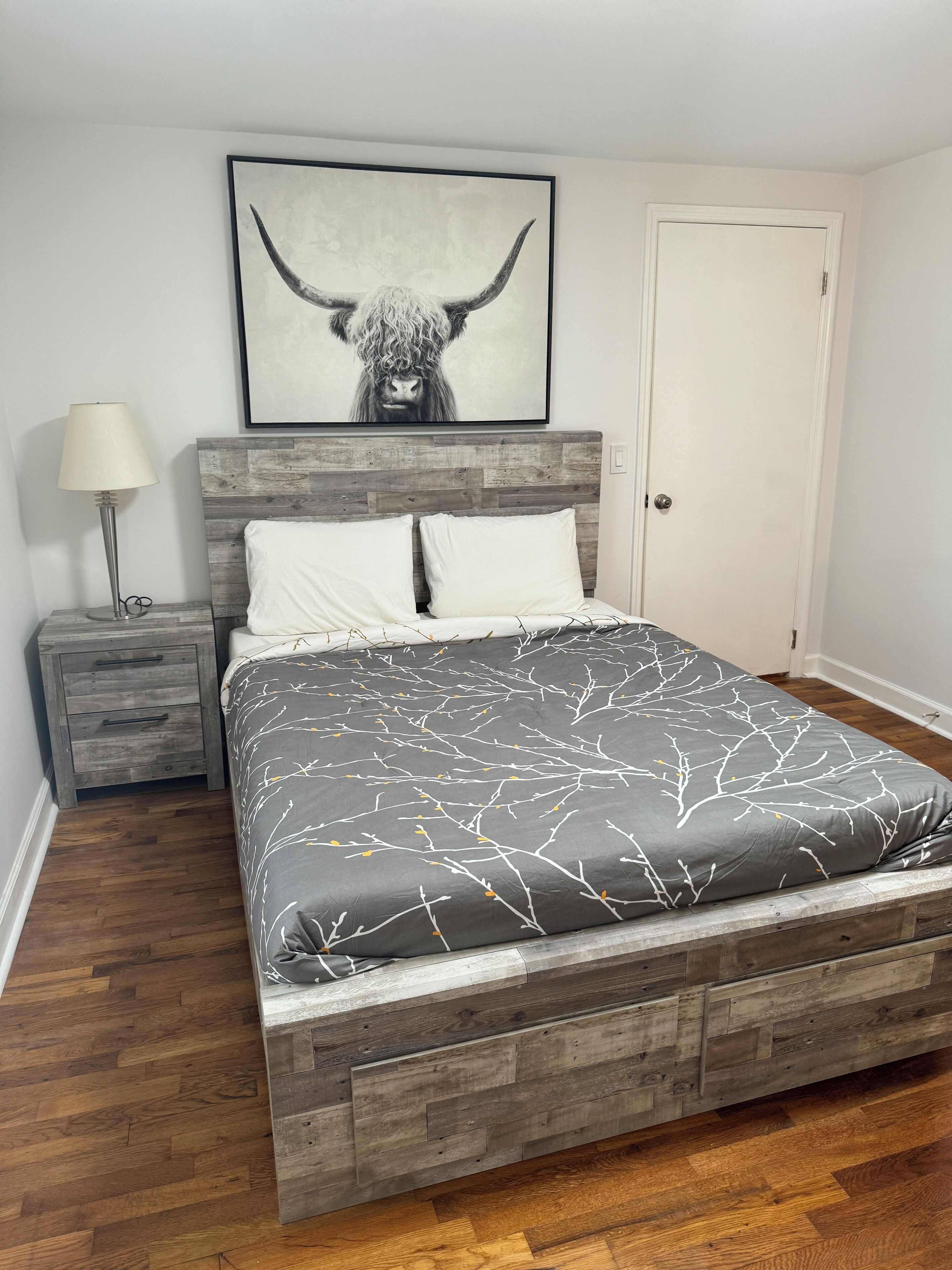 A bed with a gray and white patterned bedspread, a wooden headboard, and a framed artwork of a bull's head above it.