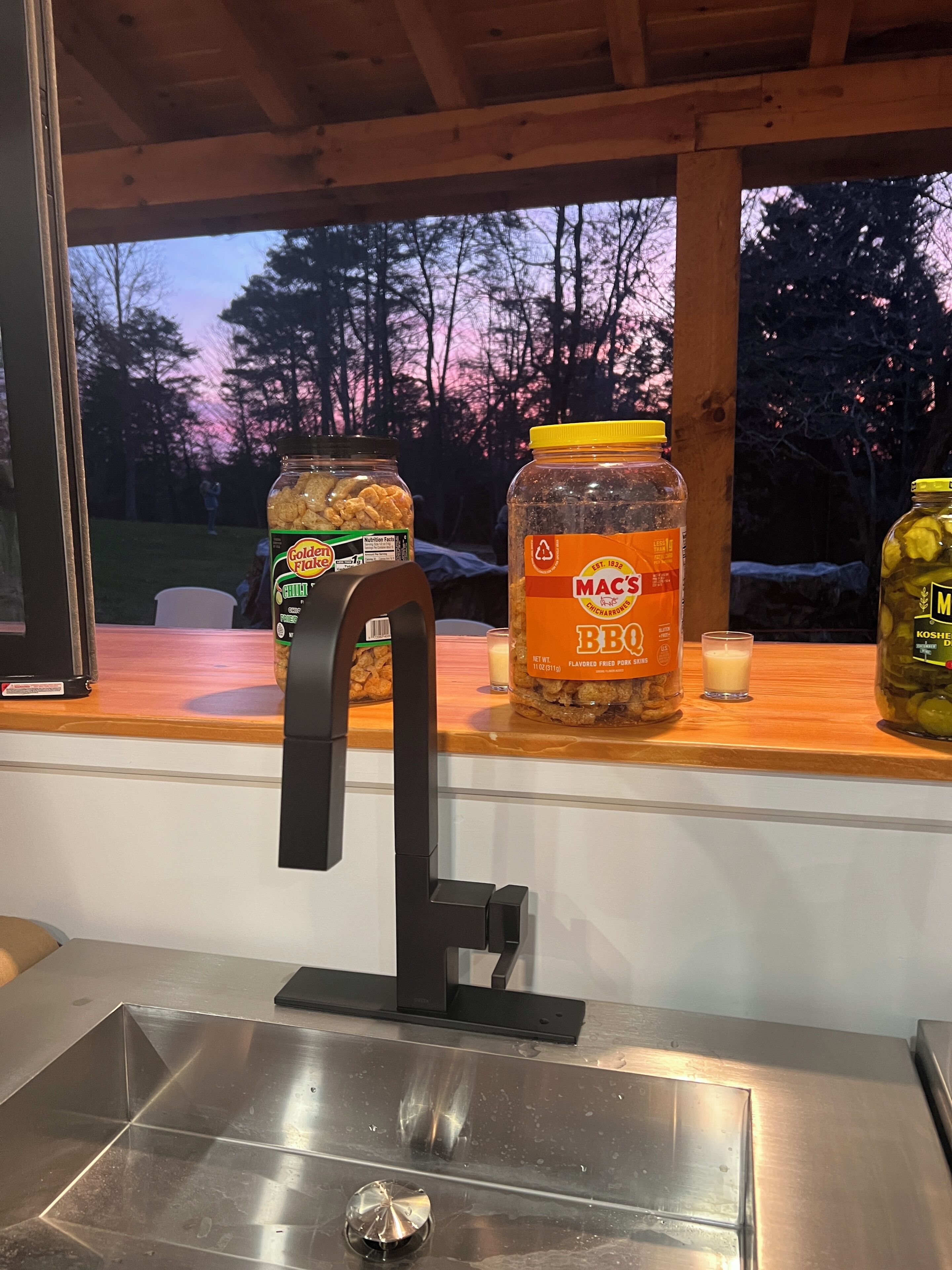 Een keukentrap met een roestvrijstalen spoelbak, een houten vensterbank met potten vol snacks en sauzen, en uitzicht op bomen en een zonsondergang.