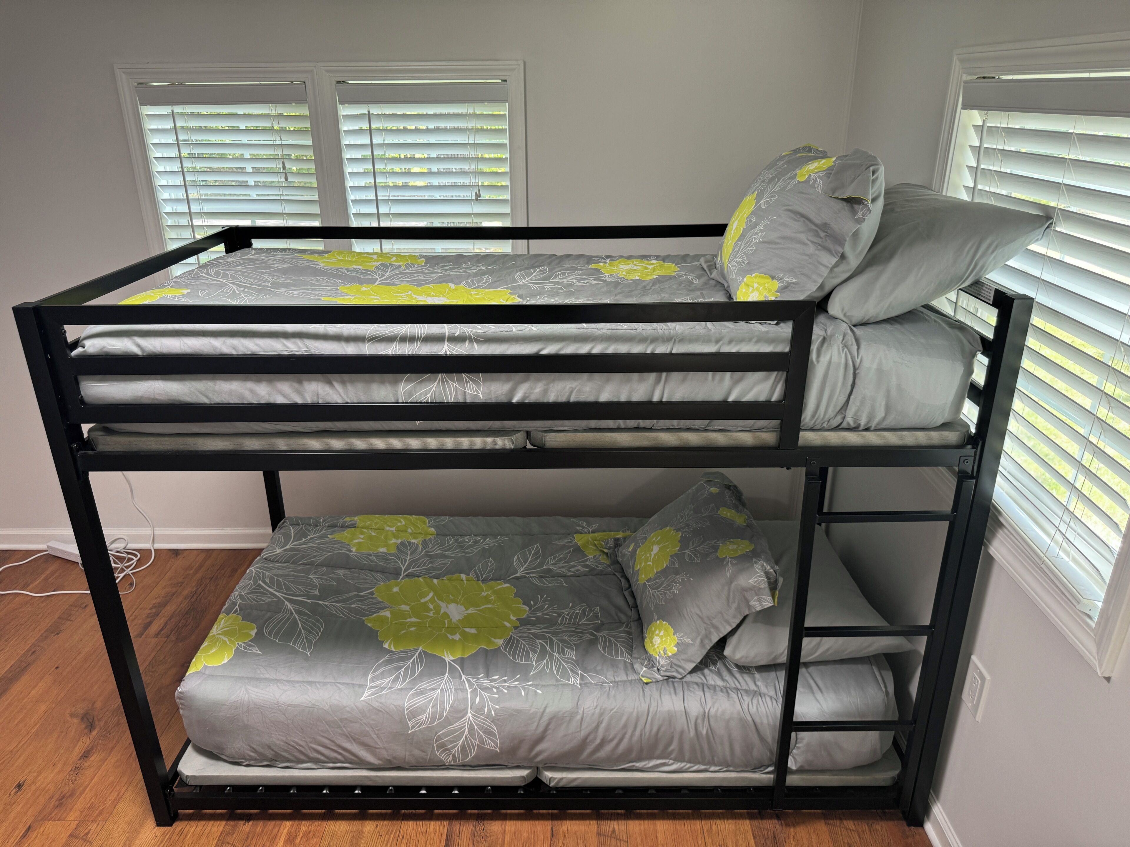 Un lit superposé avec une literie à motifs floraux gris et jaune, des stores blancs et un plancher en bois.