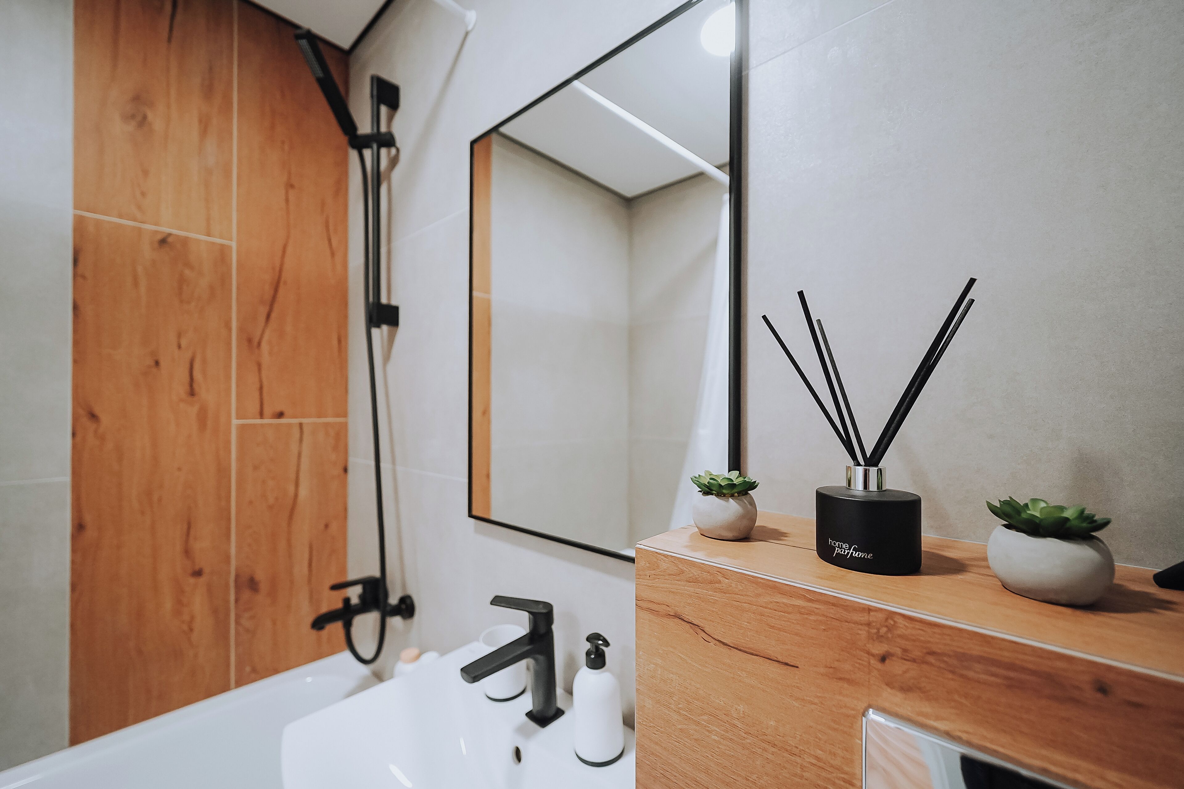 Un baño moderno con tocador de madera, un lavamanos blanco, grifo negro y un espejo. Hay dos pequeñas macetas con plantas y una botella de perfume negra sobre el tocador.