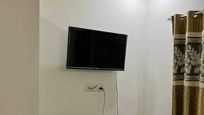 A room with a flat-screen TV mounted on the wall, a small wooden cabinet, a black plastic chair, and a patterned curtain.
