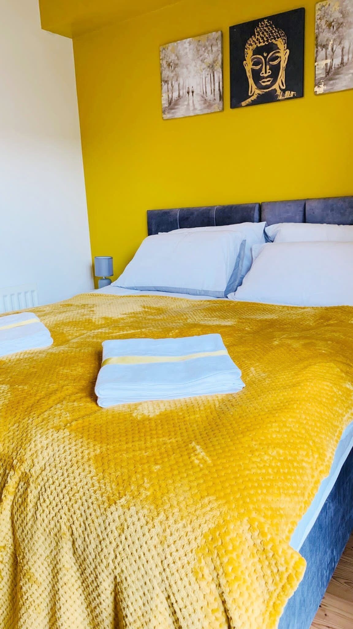 A bed with a yellow blanket and white pillows, a nightstand with a lamp, and two paintings on the wall.