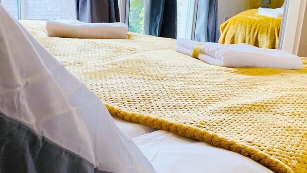 A bed with a yellow knitted blanket and white pillows, a grey pillow, and a white duvet.