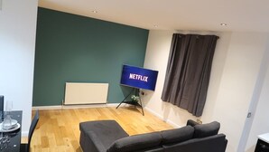 Un salón con un sofá negro, suelo de madera, televisor sobre un mueble bajo y una ventana con cortinas.